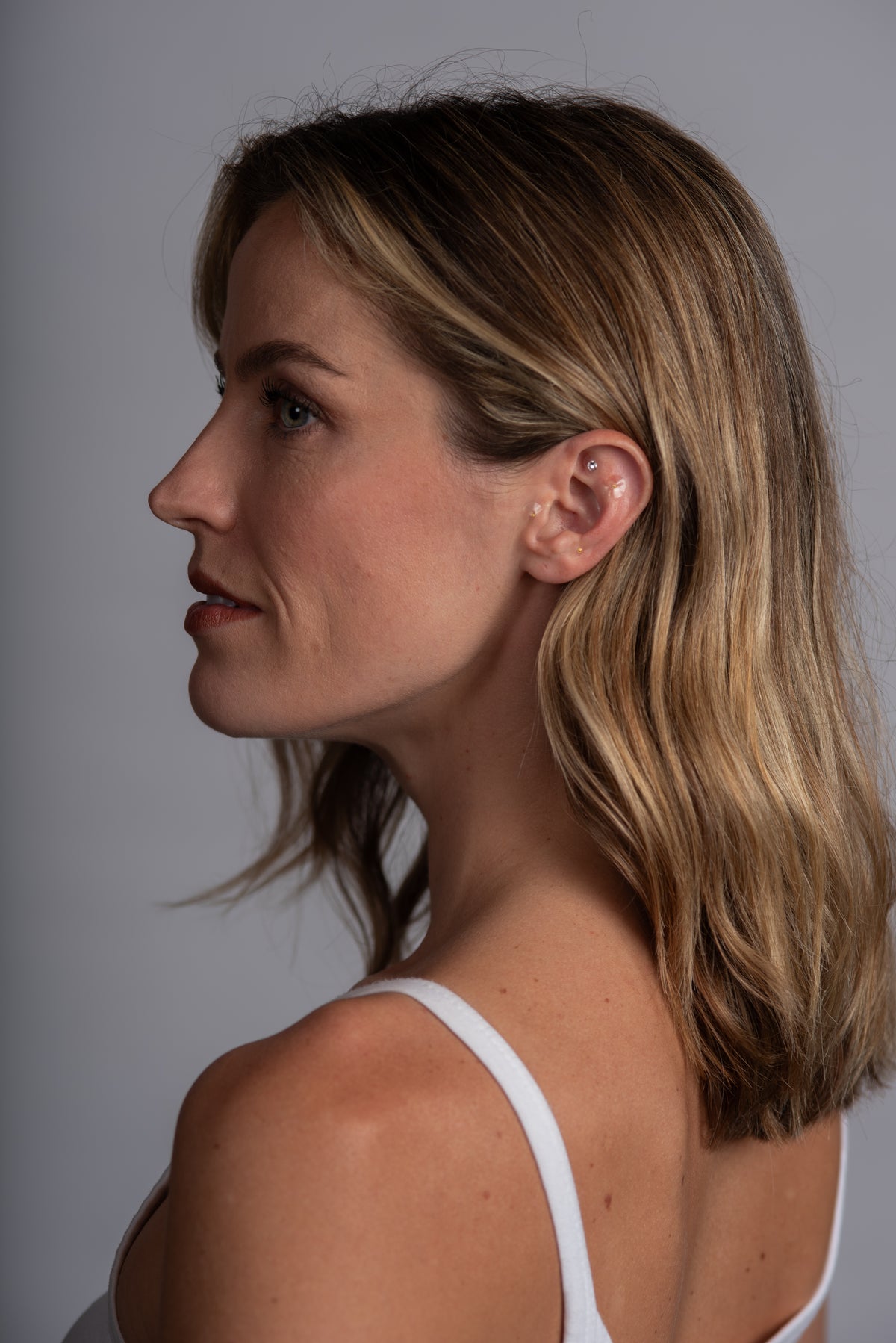 Woman wearing ear seeds in a white tank top with a grey backgound.